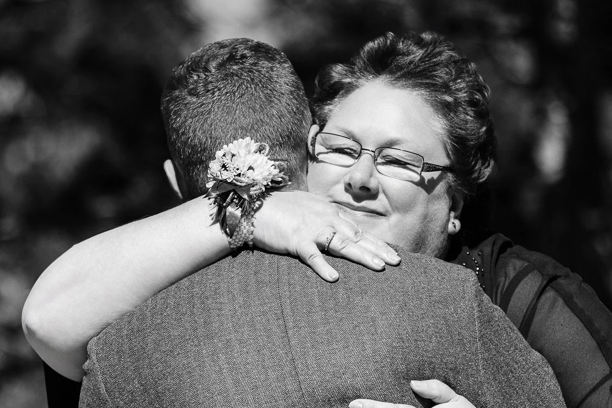 A woman with glasses gently embraces a man in a close hug, smiling warmly. Her wrist has a floral corsage. The scene conveys warmth and affection.