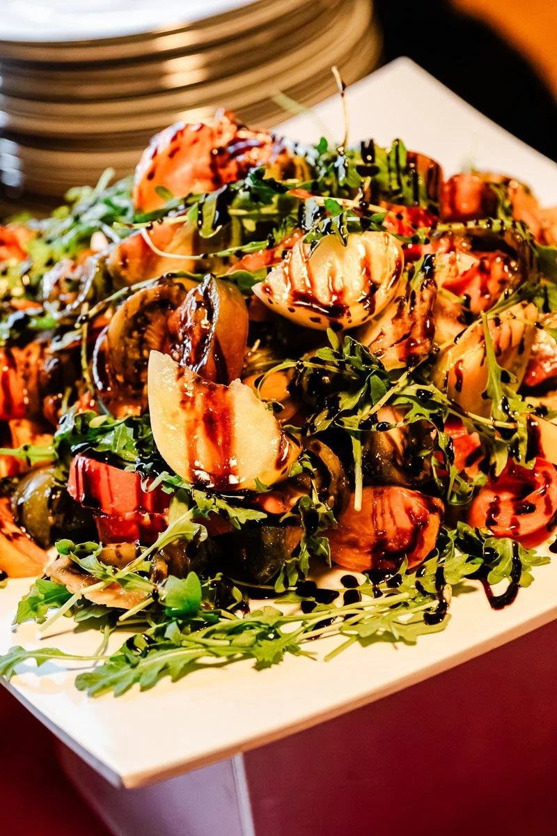 A vibrant salad with sliced tomatoes, arugula, and balsamic glaze on a square white plate. The setting is elegant, evoking a gourmet dining feel.