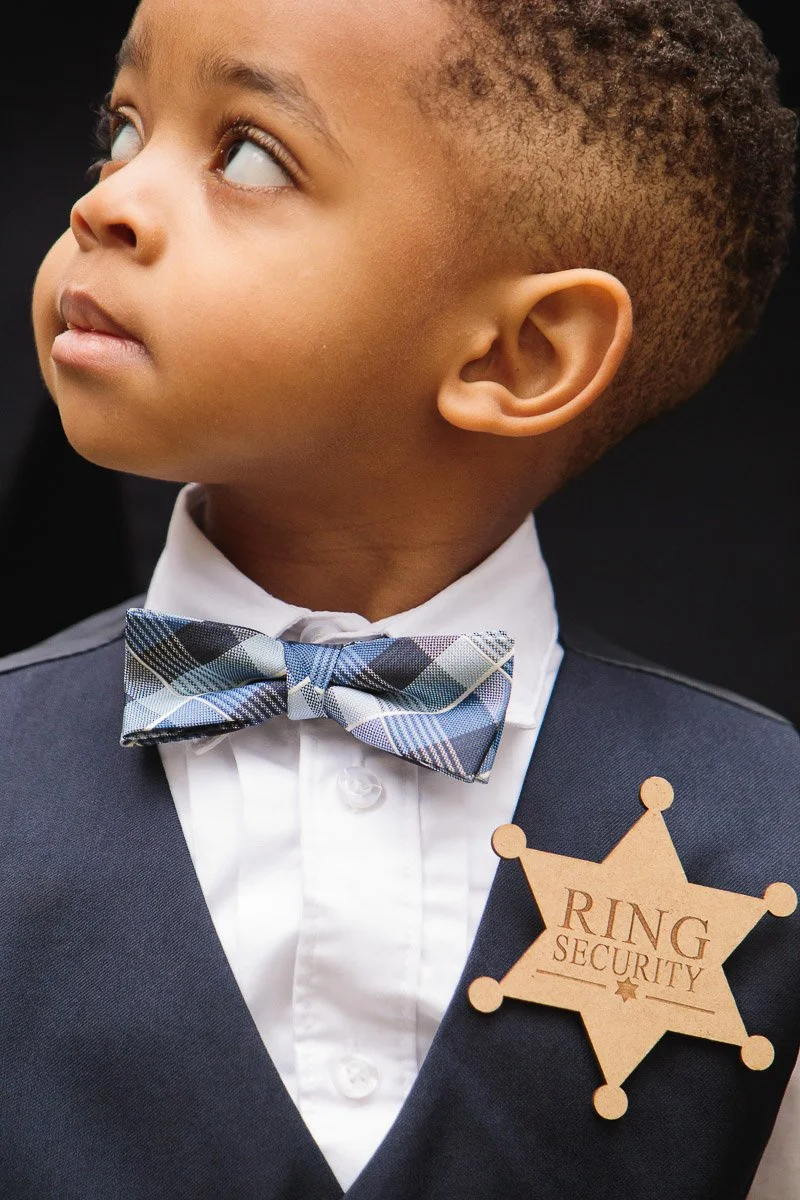 A young child looks upwards, wearing a plaid bow tie and a vest. The child has a "Ring Security" badge pinned to the vest, conveying a playful tone.