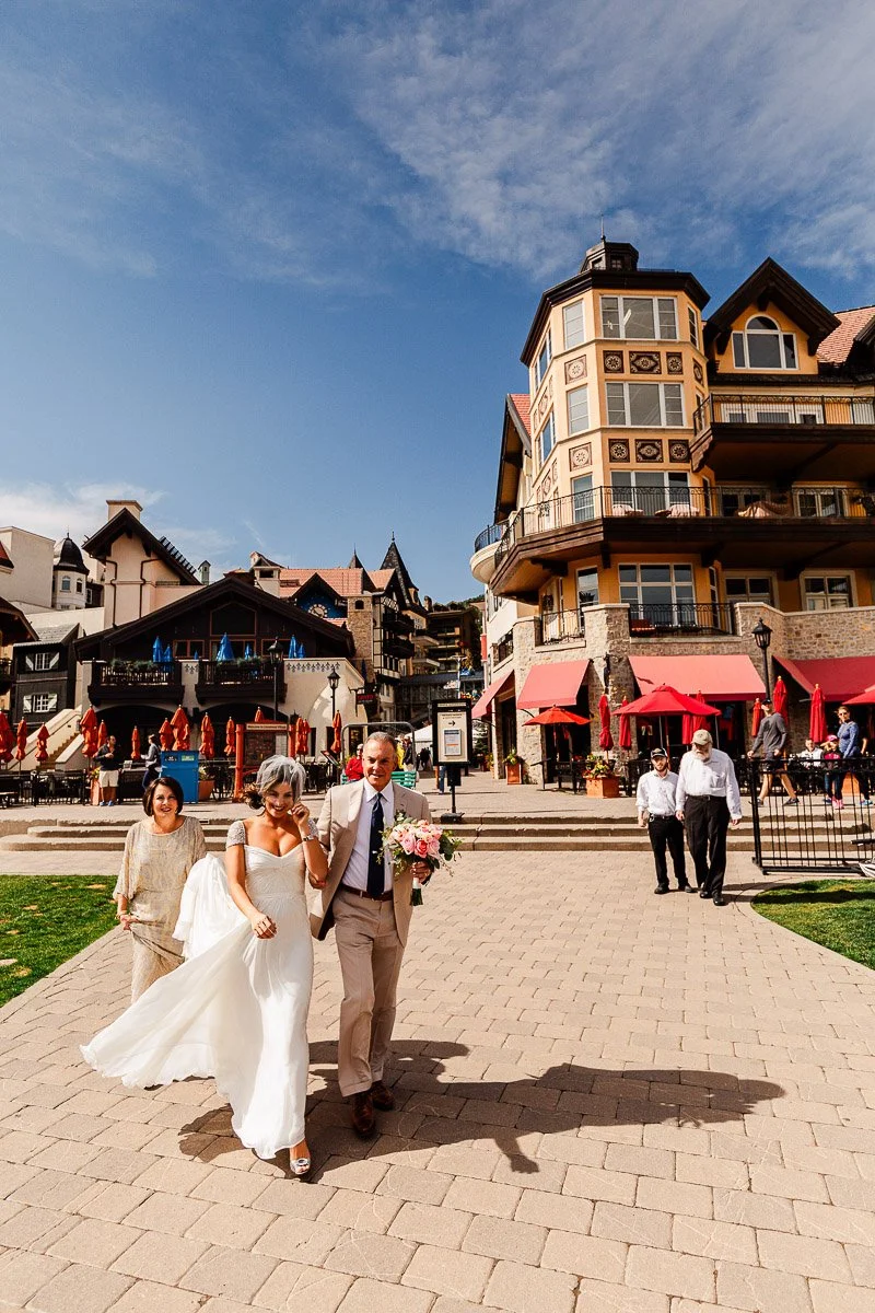 Bride in flowing white dress walks with two companions on a sunny street with European-style buildings, evoking joy and festivity.