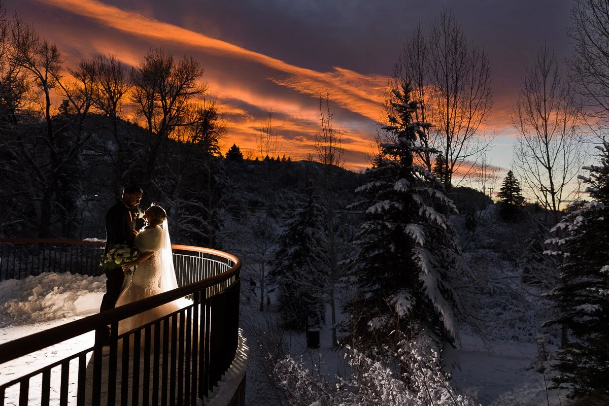 A couple embraces on a snowy bridge with a dramatic sunset backdrop, casting warm orange and purple hues over the winter landscape, conveying romance captured by Beaver Creek wedding photographer tomKphoto
