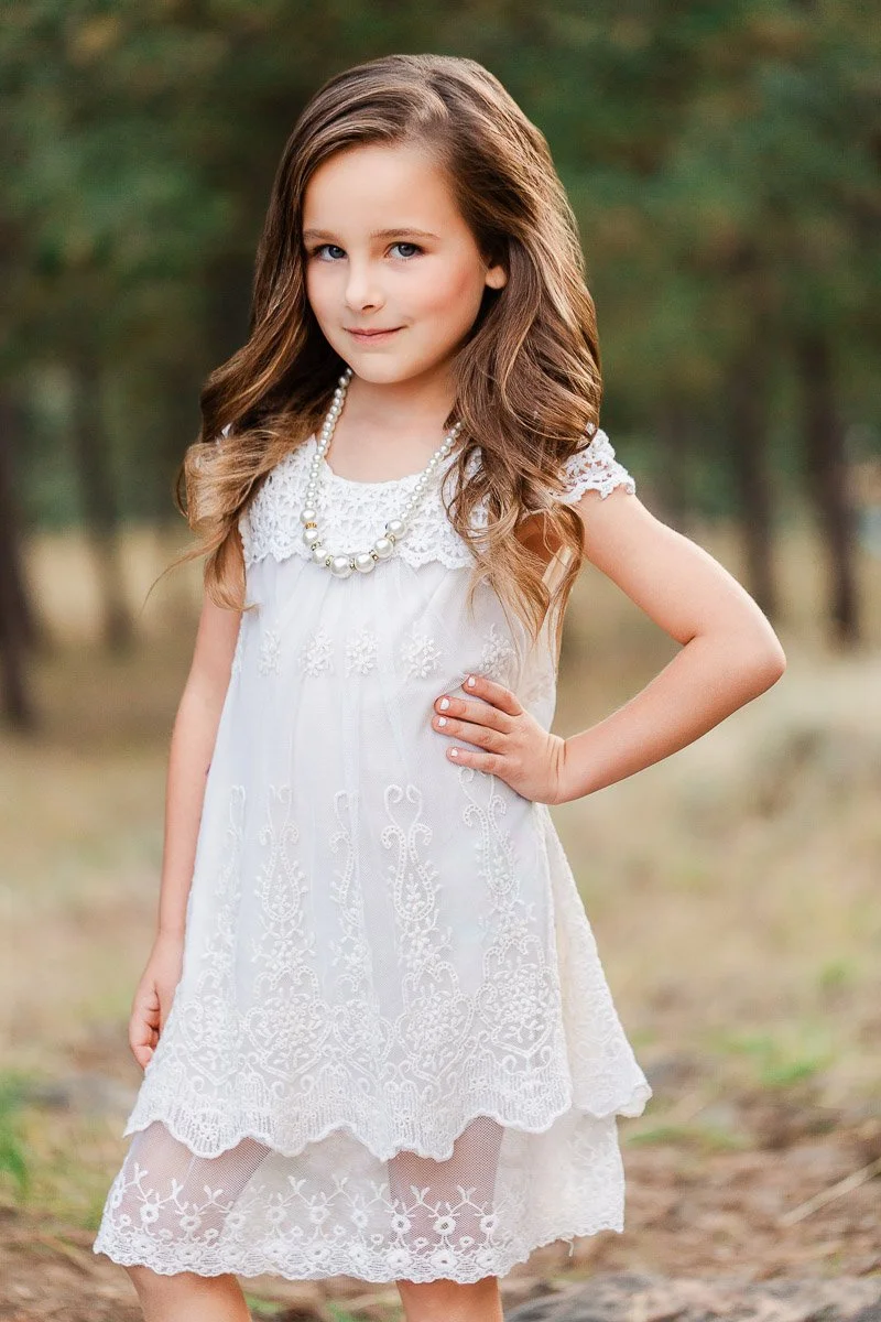 Young girl in a white lace dress with pearls stands confidently, hand on hip, in a forest. Soft focus background creates a serene, dreamy atmosphere.