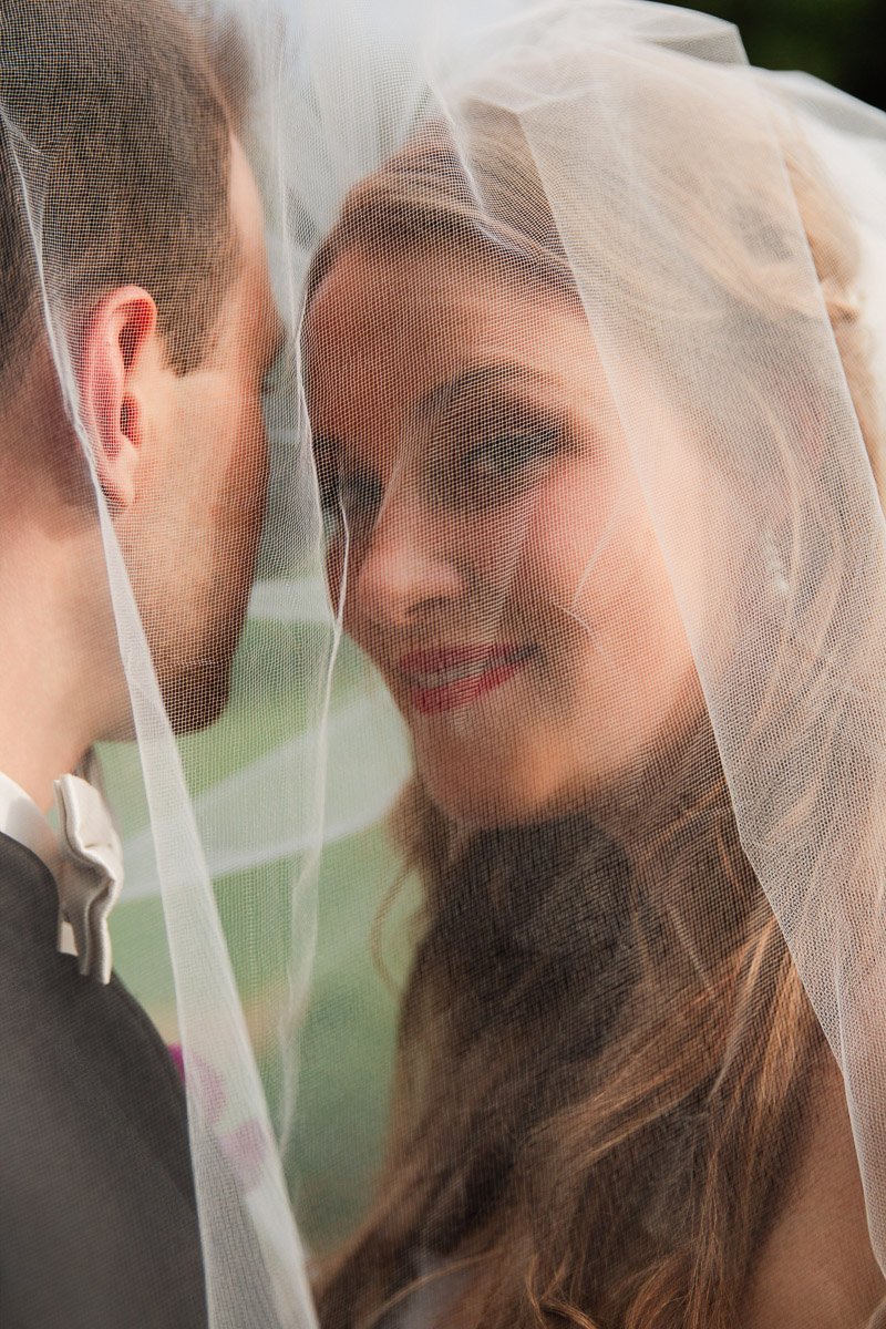 Bride and groom under a veil, close and smiling. The bride looks at the groom affectionately, radiating happiness. Soft, romantic atmosphere.
