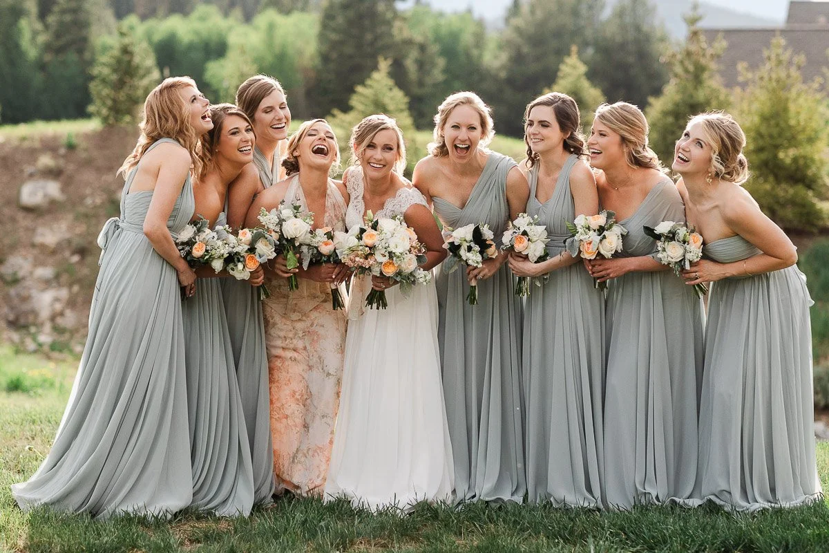A joyful group of bridesmaids in pale blue dresses and a bride in white stand outdoors, holding bouquets and laughing together in a scenic, wooded setting captured by Breckenridge wedding photographer tomKphoto
