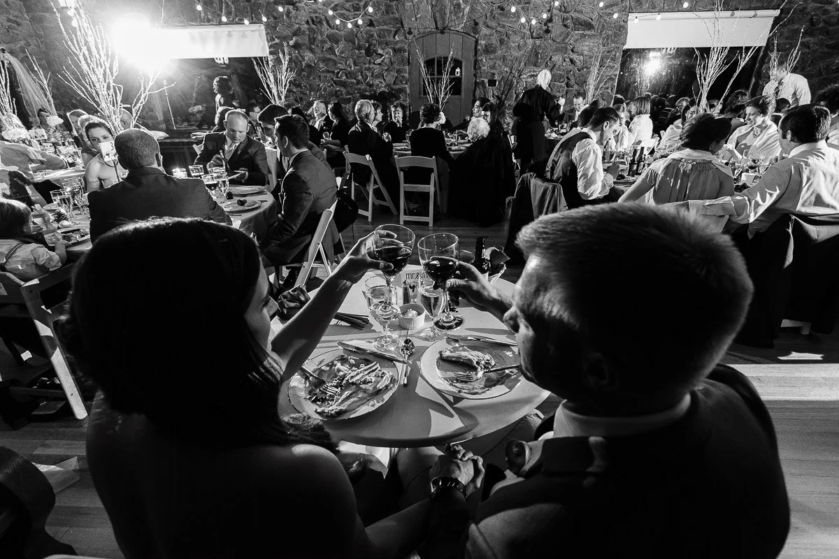A couple in formal attire toasts with wine glasses at a bustling, elegant event. Warm lighting and decor create a festive, joyful ambiance.