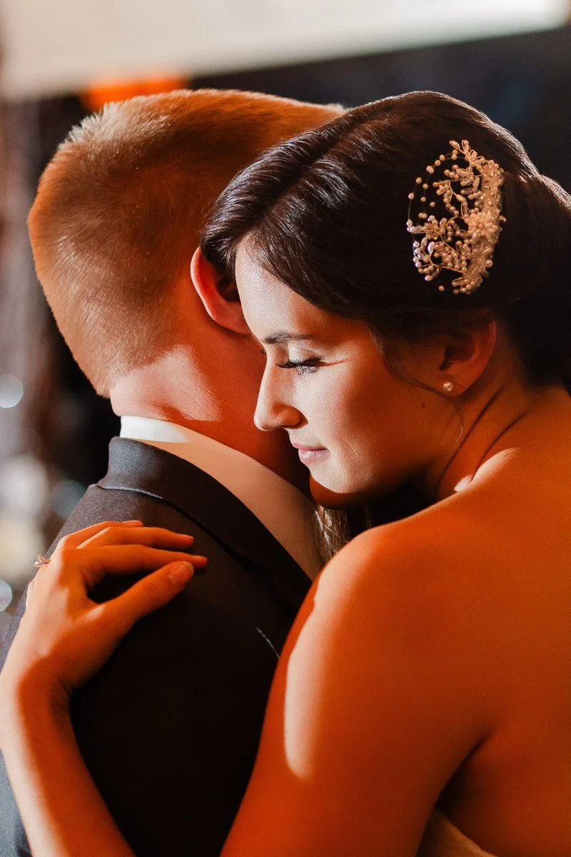 A couple embraces closely, the woman in a wedding dress with a decorative hair accessory, eyes gently closed, conveying warmth and intimacy.