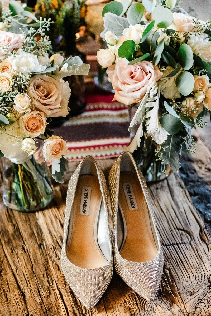 Elegant glittery pumps placed on rustic wooden surface, flanked by lush bouquets of pink roses and greenery, creating a romantic, vintage ambiance.
