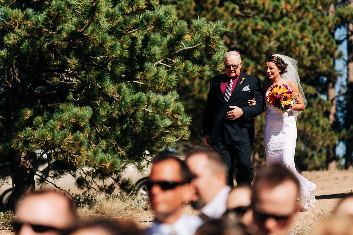 Bride in white gown and holding colorful bouquet walks arm-in-arm with man in suit under sunny skies, surrounded by pine trees and guests.