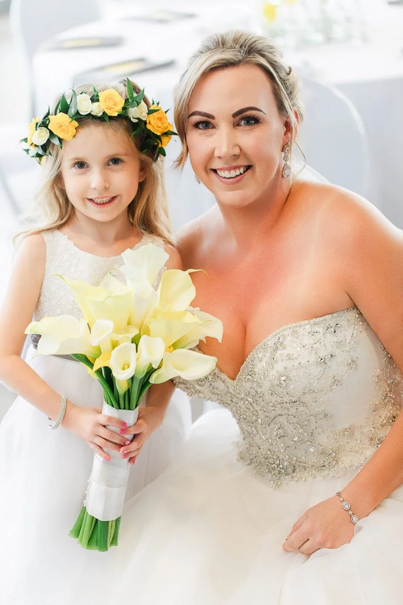 A bride with an embroidered gown smiles beside a young girl in white, wearing a floral crown. Both hold yellow calla lilies, exuding joy and warmth.