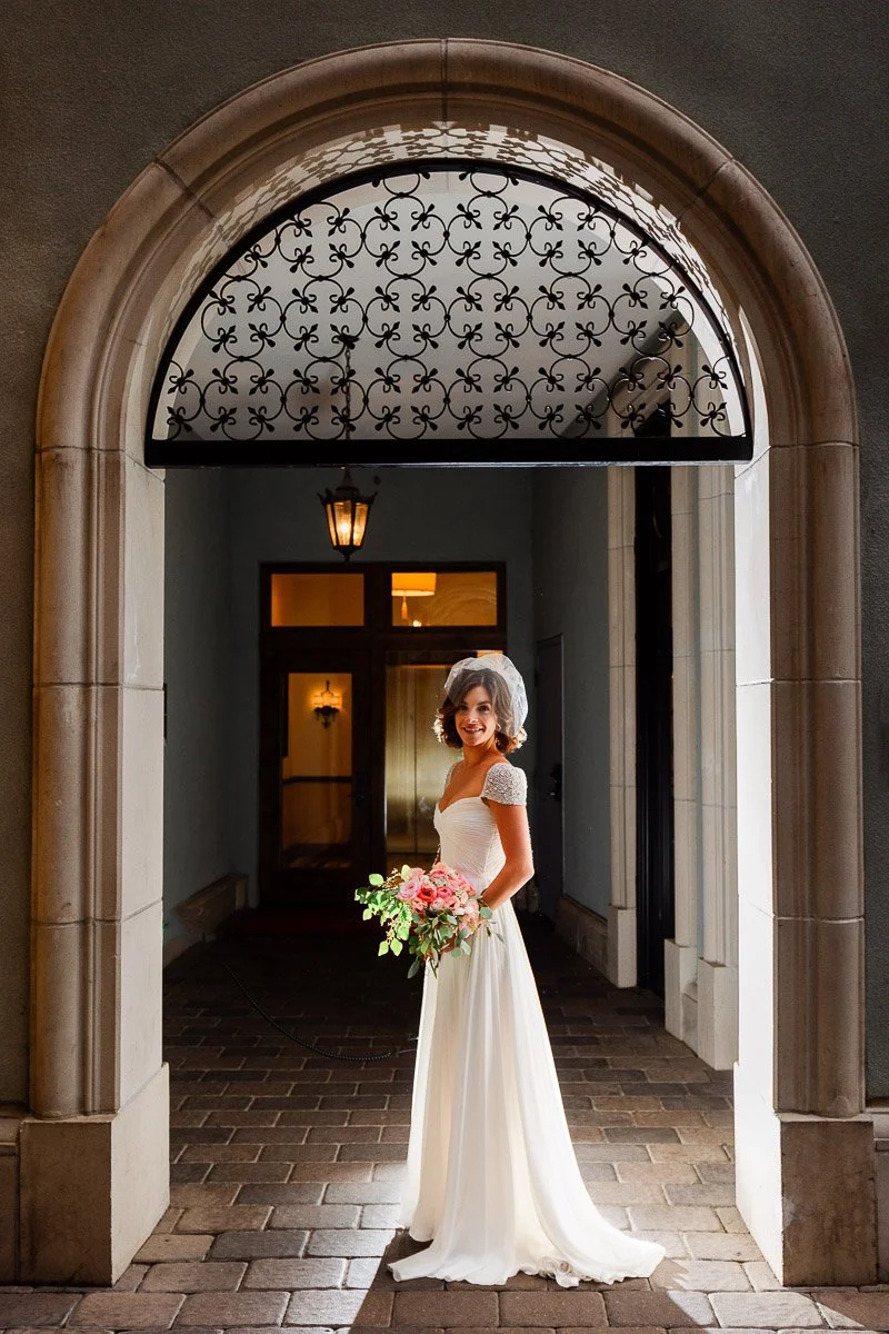 A bride in a white gown and veil stands under an ornate archway, holding a pink bouquet. Sunlight streams in, creating a warm and elegant atmosphere.