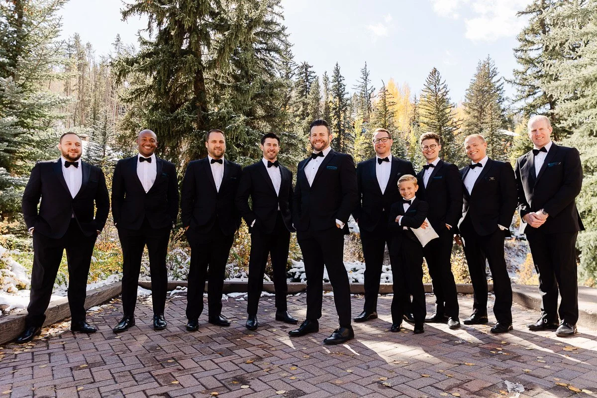 A group of nine men and a child in suits and bow ties stand confidently on a snowy pathway, surrounded by tall fir trees and autumn foliage.