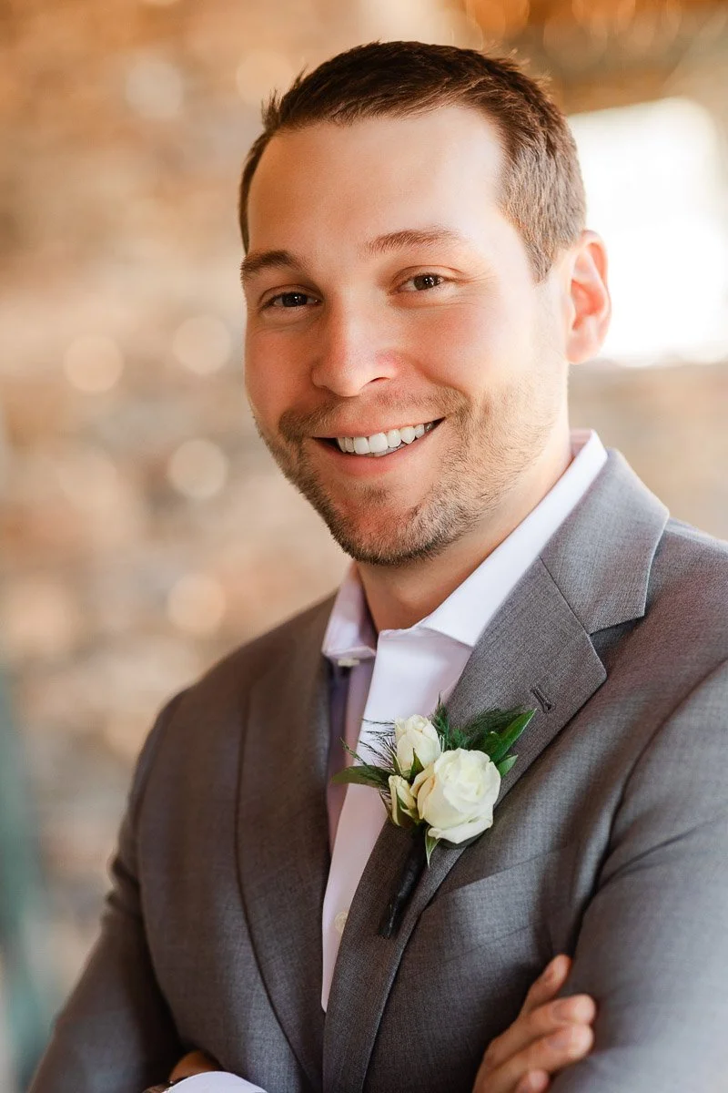 Smiling man in a gray suit with a white rose boutonniere, arms crossed. Background features a softly blurred, warm-toned interior wall.