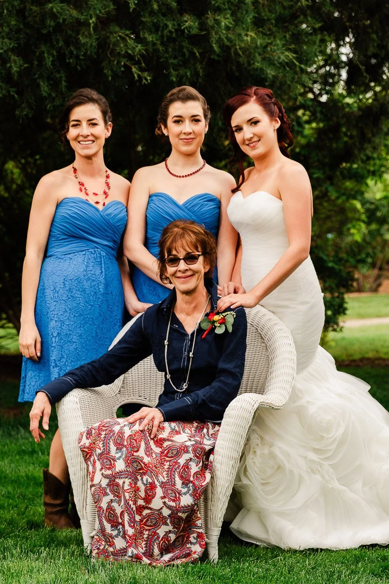 A seated woman in sunglasses and floral skirt is in front, surrounded by three standing women in blue dresses and a white gown, outdoors, smiling warmly.
