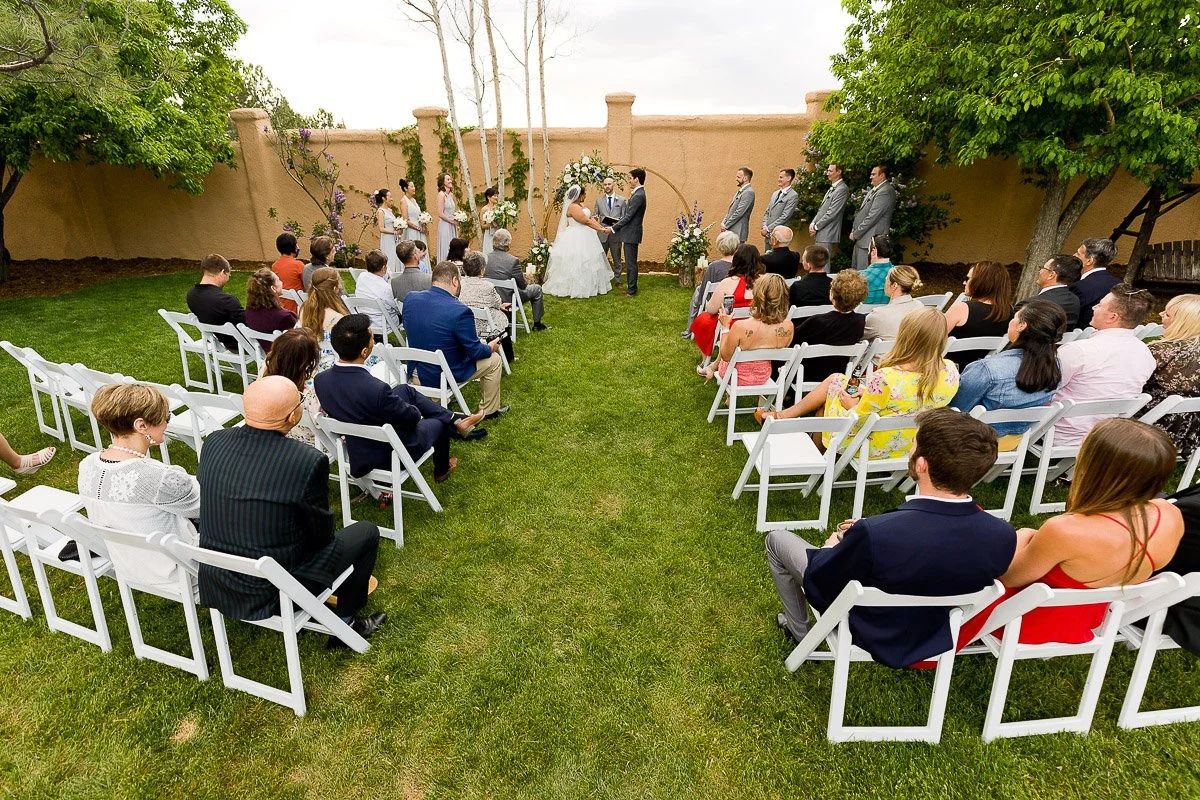 A wedding ceremony outdoors with seated guests on white chairs facing a couple under a floral arch. Bridesmaids and groomsmen stand nearby, wearing suits and dresses.