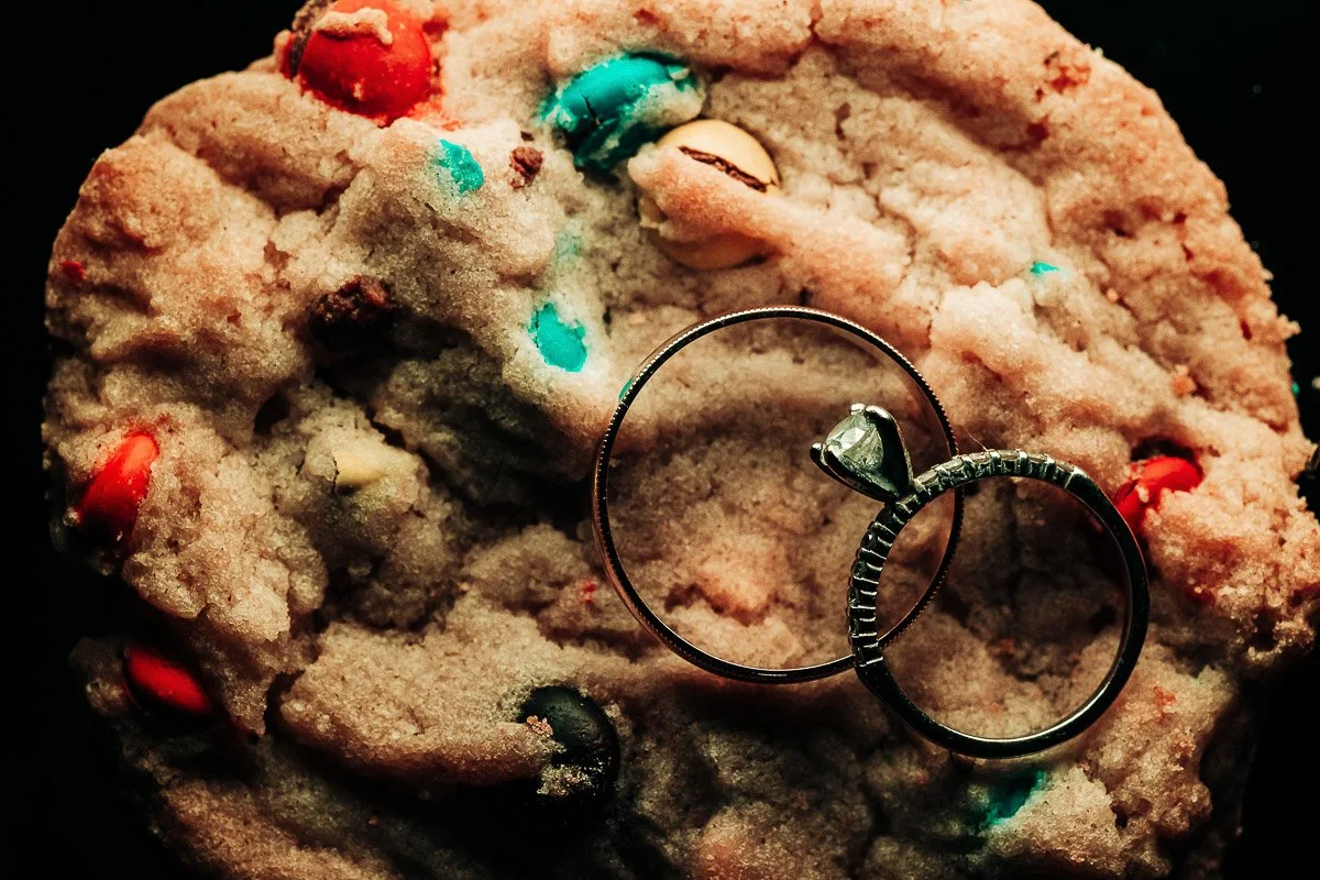 A colorful cookie studded with candies, topped by two silver rings, one with a diamond. The image conveys warmth and a touch of romance.