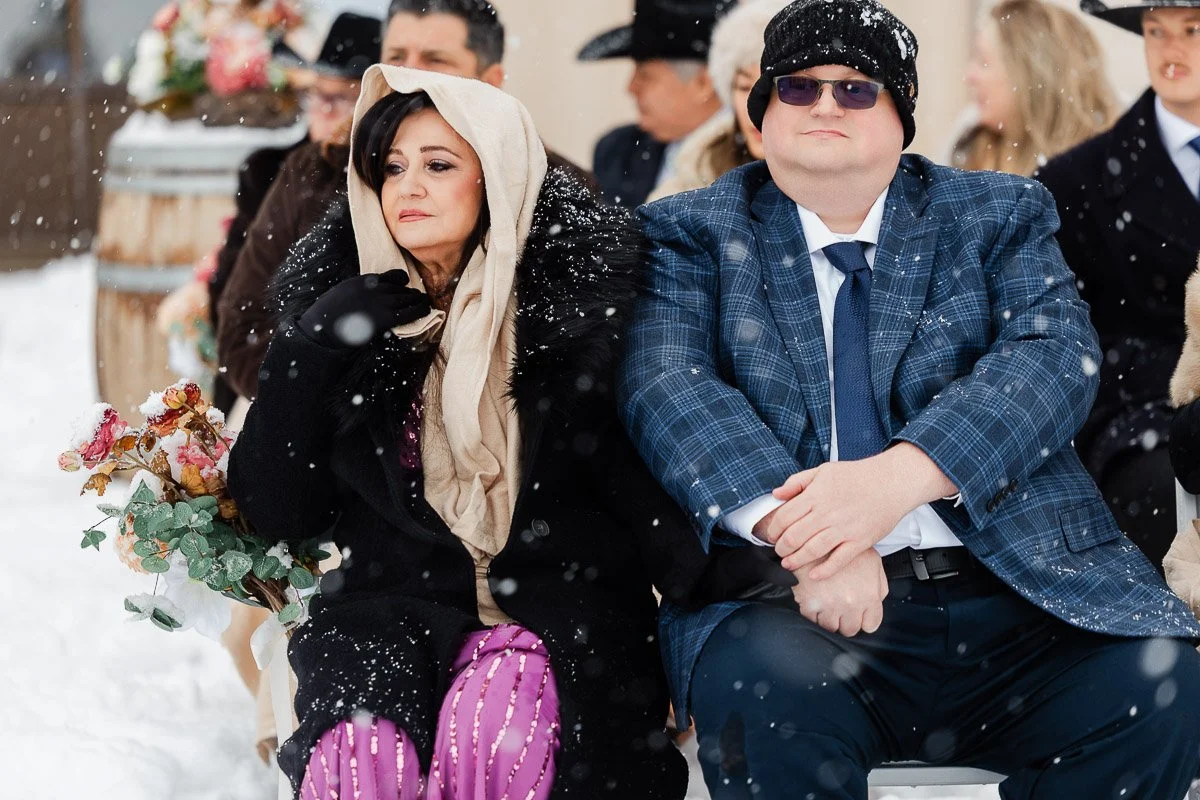 A woman in a fur coat and scarf sits next to a man in a blue suit and hat at an outdoor snowy event. They look bundled and slightly cold.