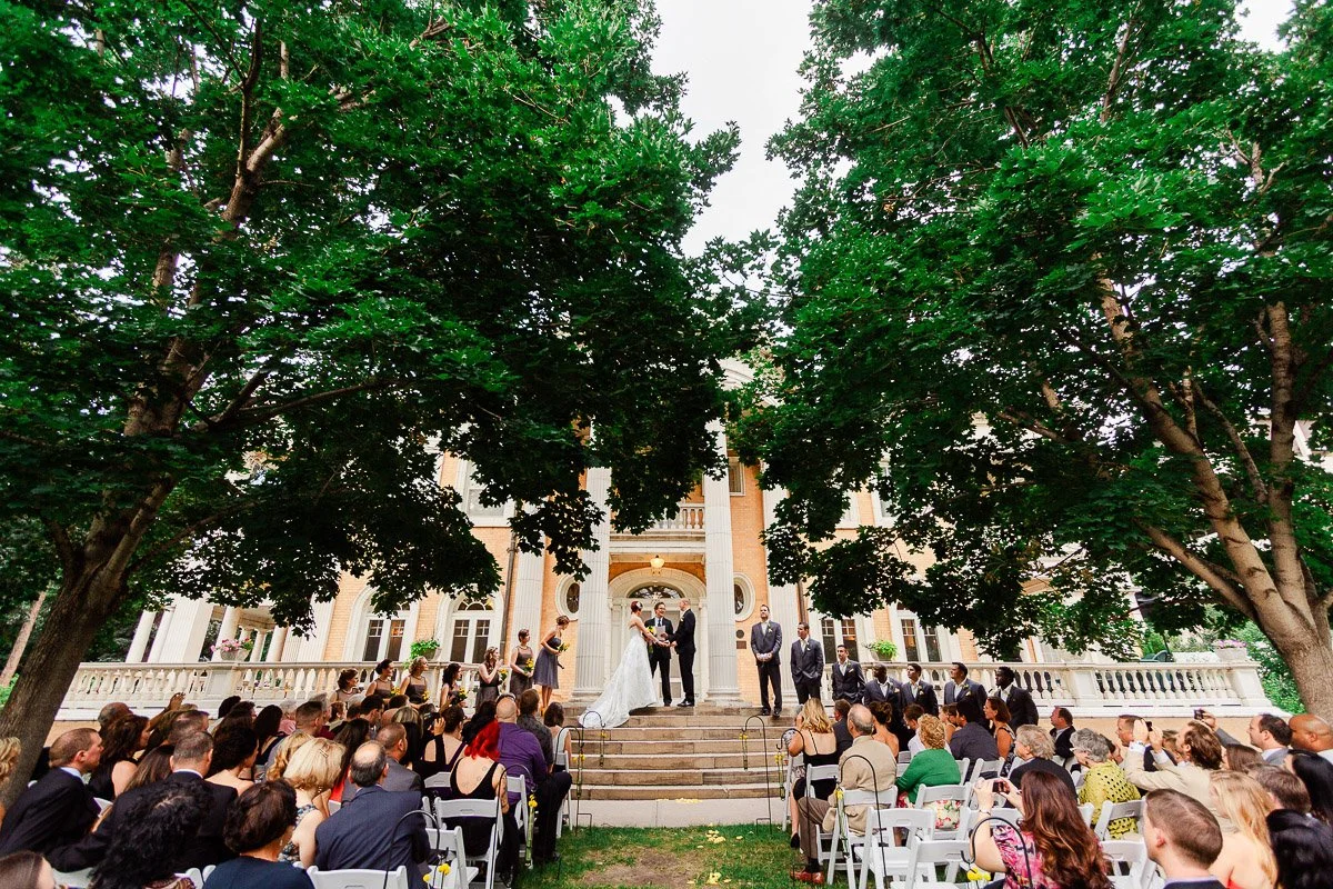 Wedding ceremony outdoors with guests seated in white chairs facing a couple under a large tree. The backdrop is an elegant mansion entrance.