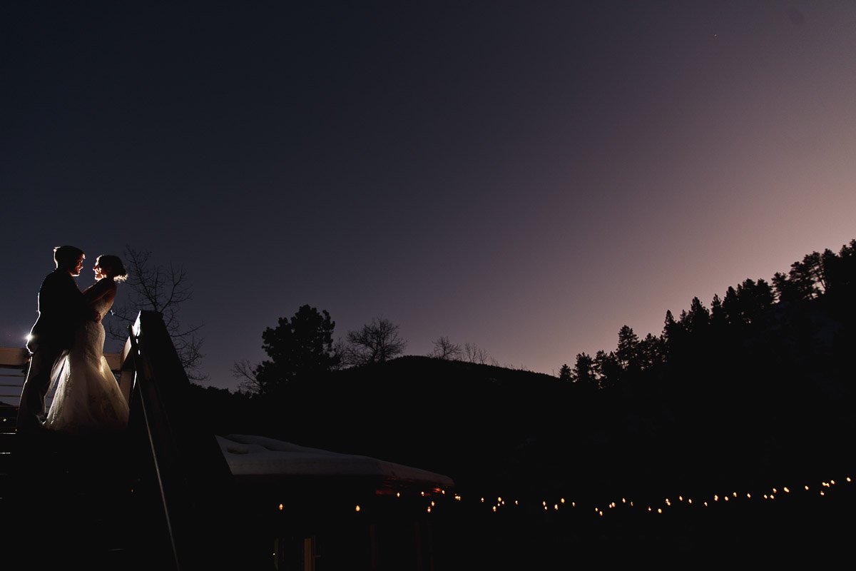 Silhouetted bride and groom embrace on a staircase at dusk, surrounded by trees and a string of warm lights, creating a romantic, serene atmosphere.