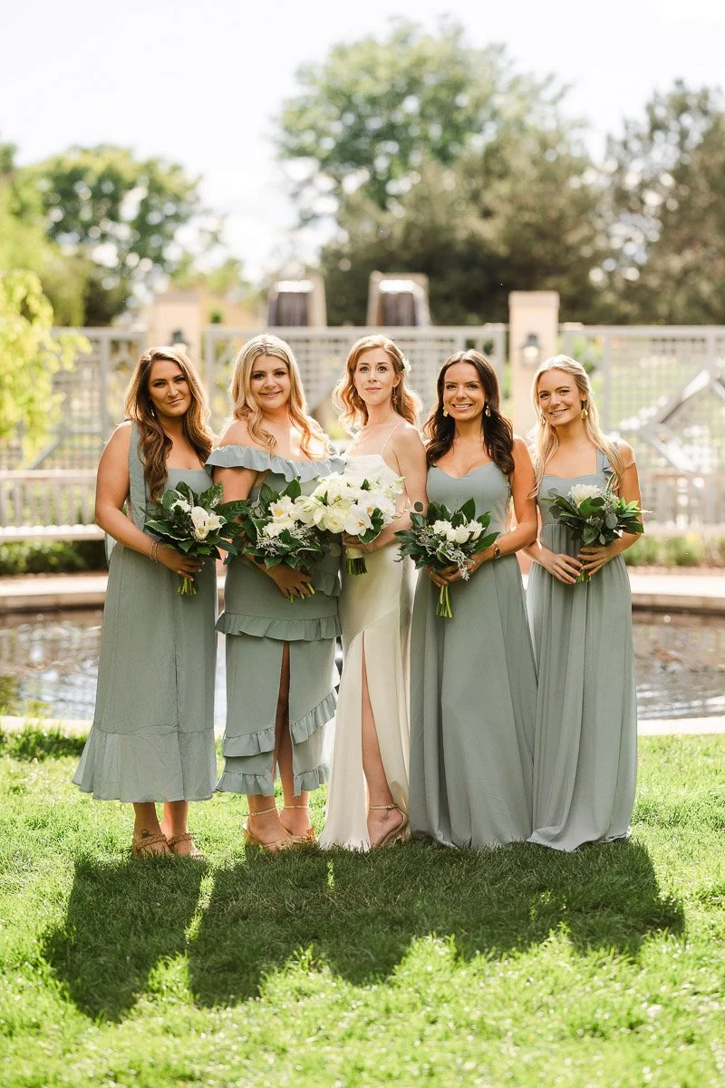A bride in a white dress stands with four bridesmaids in sage green dresses, holding bouquets, smiling in a garden setting with a sunny backdrop.