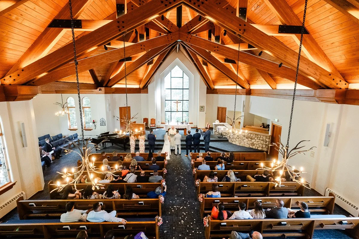 A wooden-beamed chapel interior during a wedding ceremony. The bride and groom stand before the altar, surrounded by guests seated in pews. Warm lighting and floral decorations create an intimate atmosphere.
