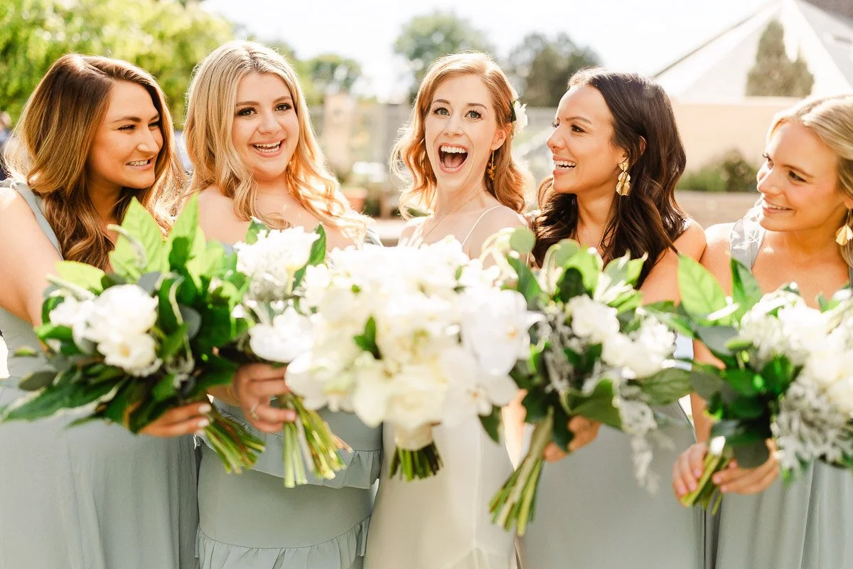 A joyful bride and four bridesmaids in light-blue dresses smile widely, holding white bouquets. The sunny outdoor background adds a vibrant, celebratory mood.