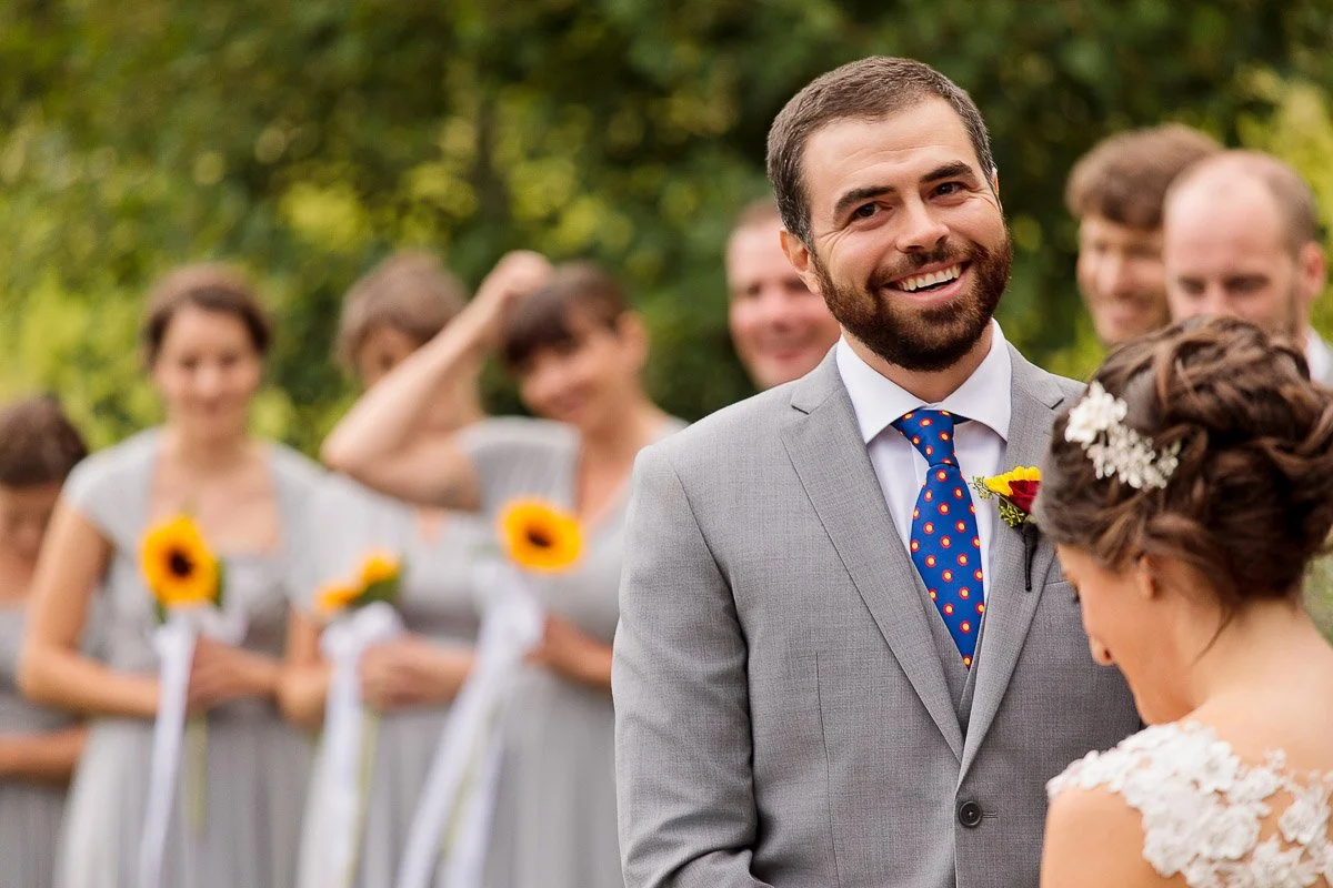 A groom in a gray suit smiles warmly at the bride, wearing a lace dress, during an outdoor wedding. Bridesmaids in gray hold sunflowers in the background.