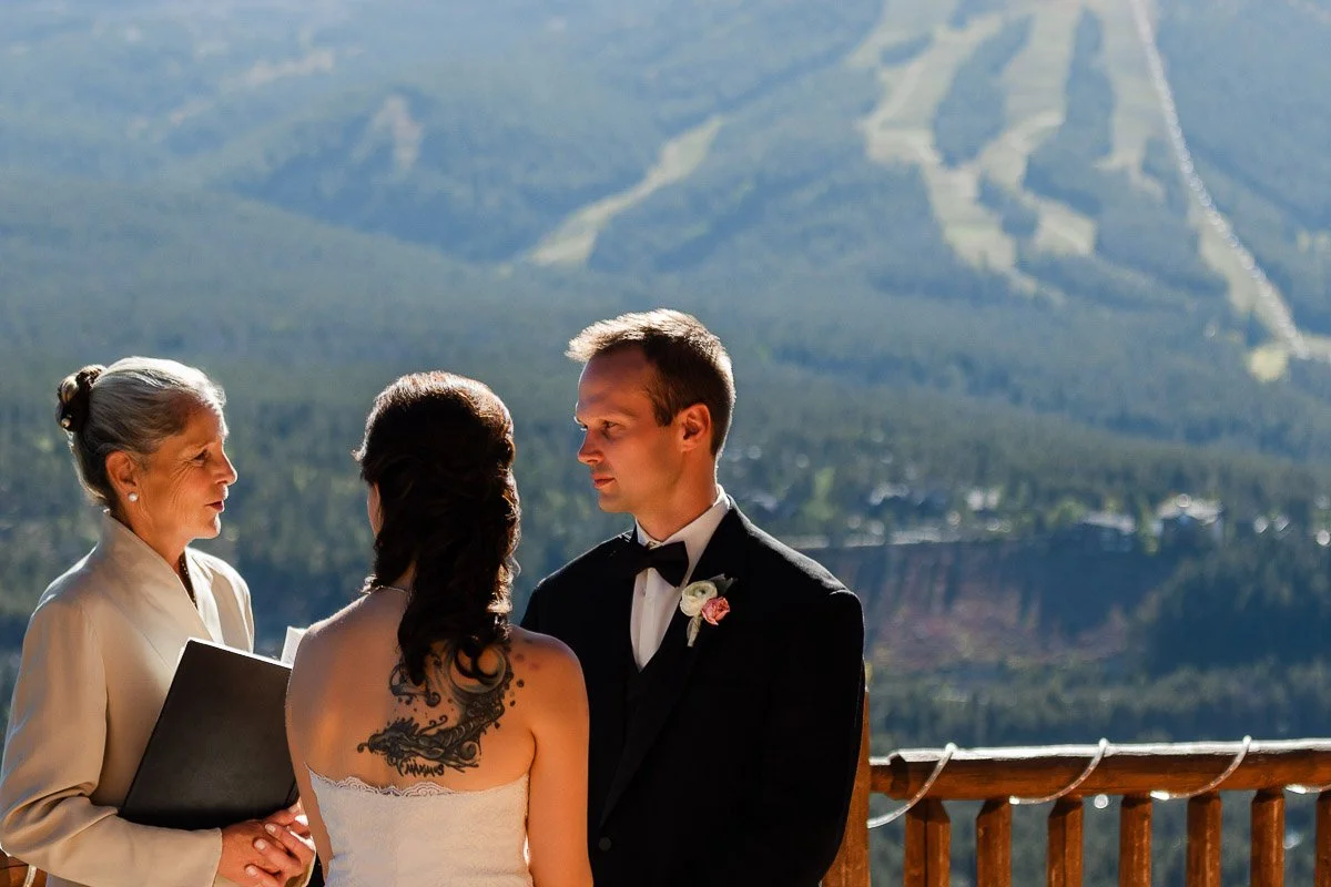 A couple stands with an officiant during an outdoor wedding ceremony. The bride has a large tattoo on her back and the groom wears a tuxedo. They're on a wooden deck with a scenic mountain view in the background.