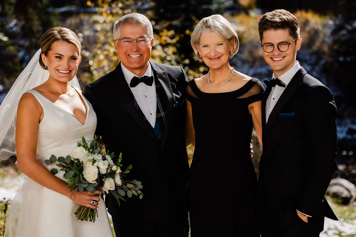 A bride and groom in elegant attire pose outdoors with two older adults, likely family. They all smile warmly in the sunlight, surrounded by autumn foliage.