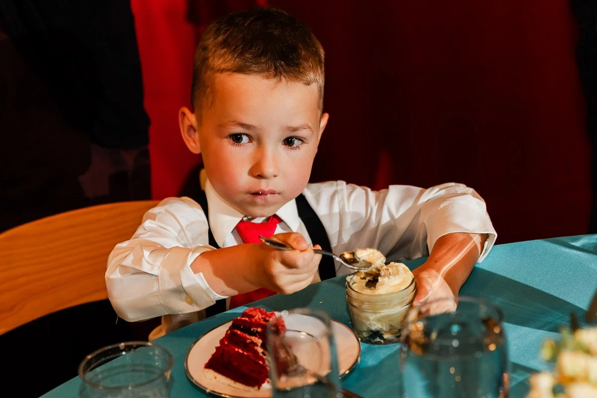 Young boy in a formal outfit enjoys dessert at a dining table. He has a focused expression, holding a spoon with a jar of food and a plate with cake.