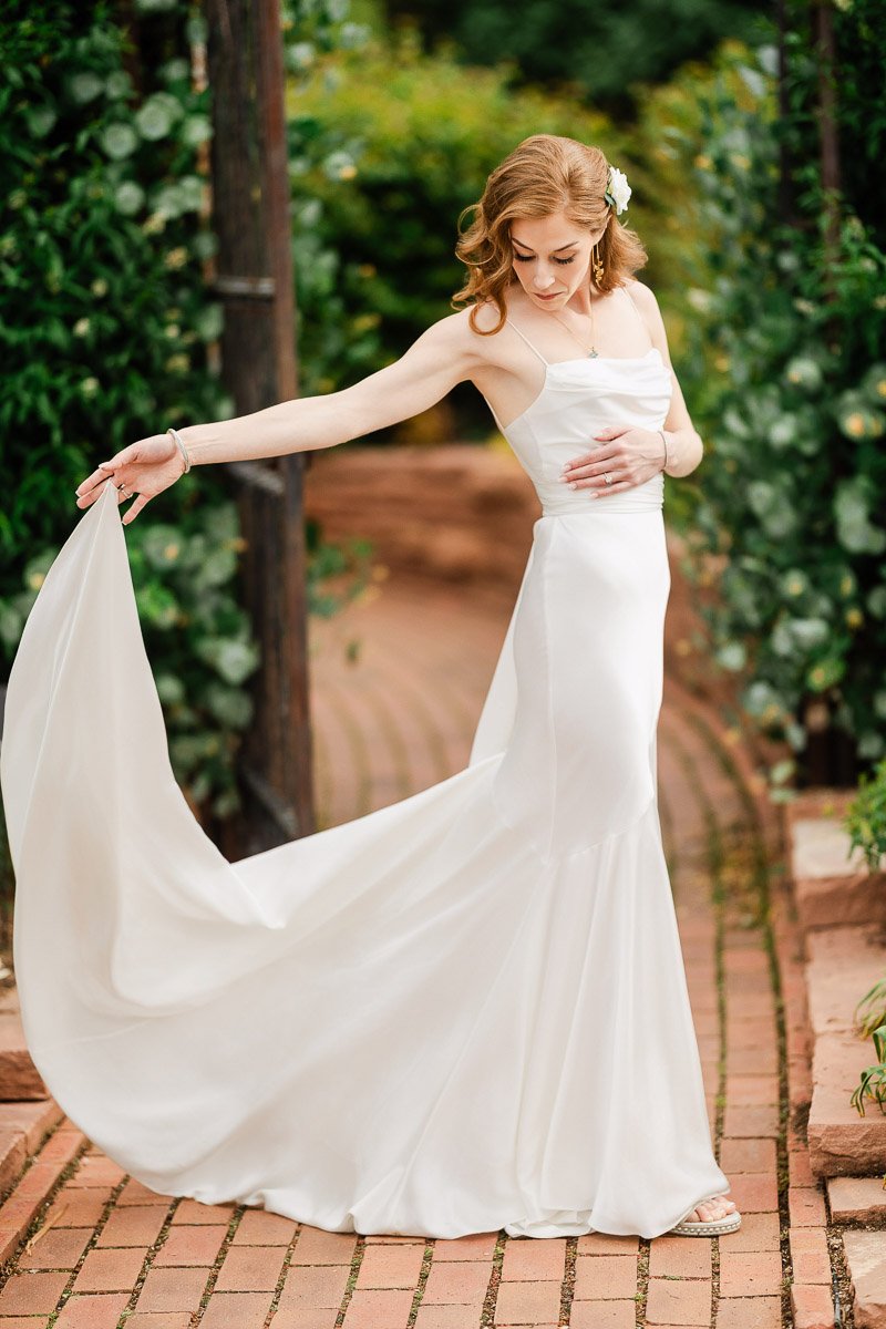 A woman in a white, flowing wedding dress gracefully poses on a brick path, surrounded by lush greenery. Her expression is serene and contemplative.