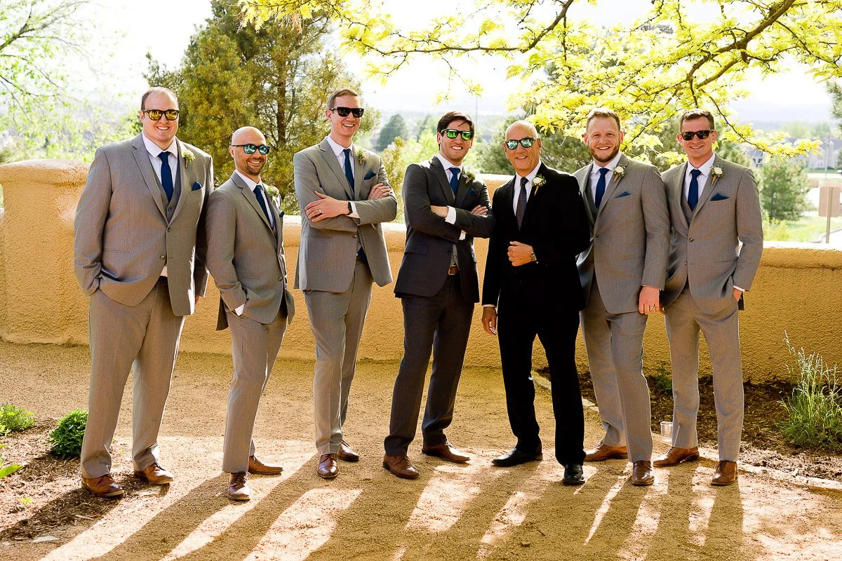 A group of seven men in suits stands outdoors on a sunny day, smiling confidently. Six wear gray suits with blue ties, and one wears a black suit.