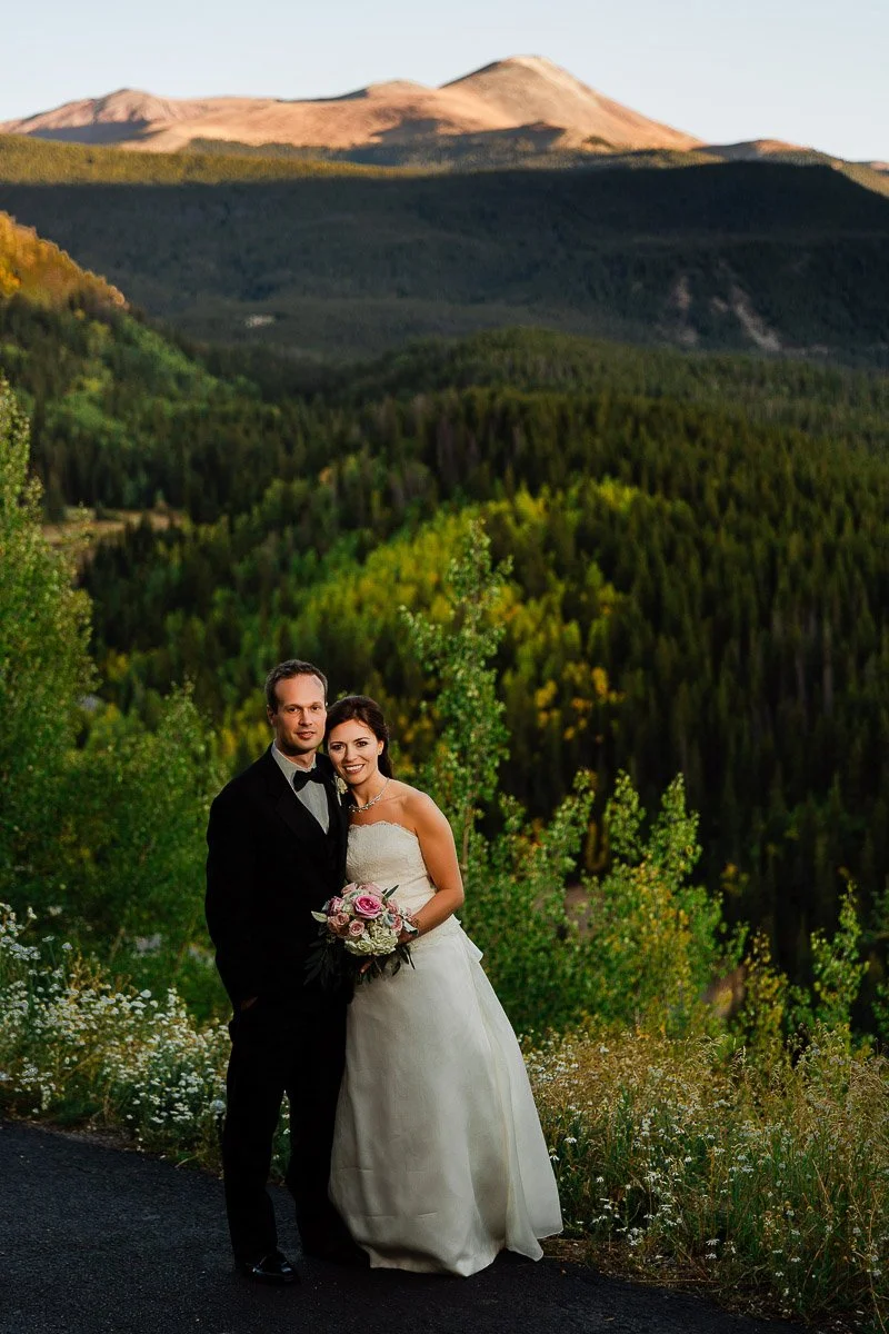 A bride and groom in formal attire stand smiling amidst lush green foliage, with a scenic mountain landscape in the background under soft lighting.