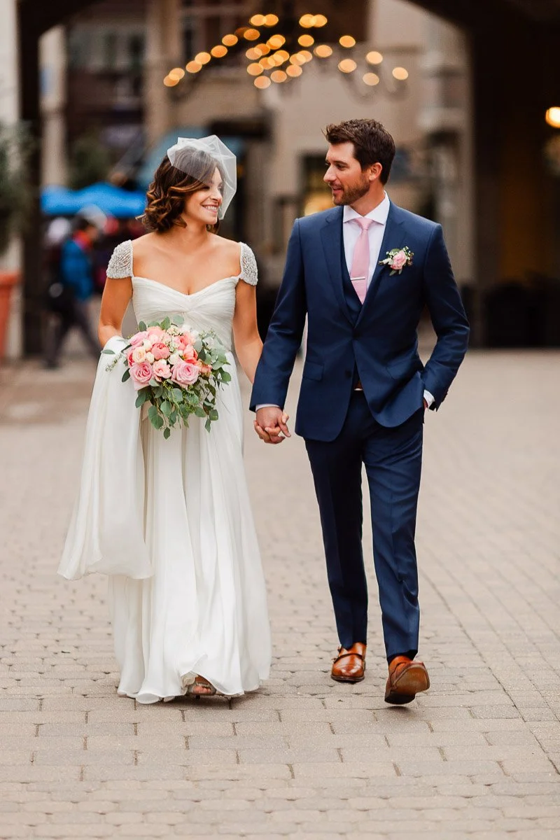 A bride in a white gown holds a pink bouquet, walking hand-in-hand with a groom in a blue suit. Both smile warmly, under soft string lights.
