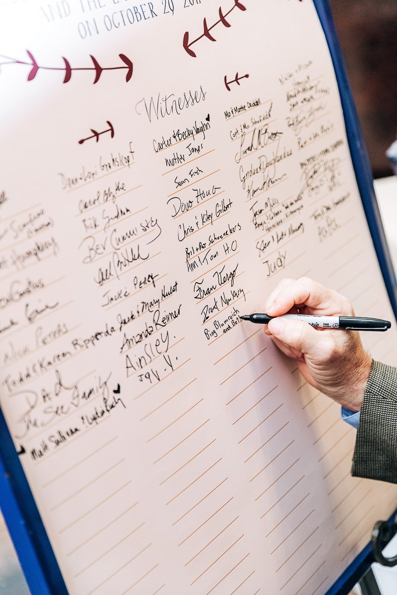 A hand signs a large guest board filled with handwritten signatures labeled "Witnesses." The board is themed with baseball stitches at the top, conveying a celebratory and formal tone.