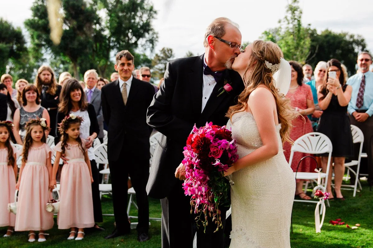 A bride in a white dress kisses a man in a tuxedo, holding vibrant pink flowers, surrounded by guests and flower girls standing on green grass.