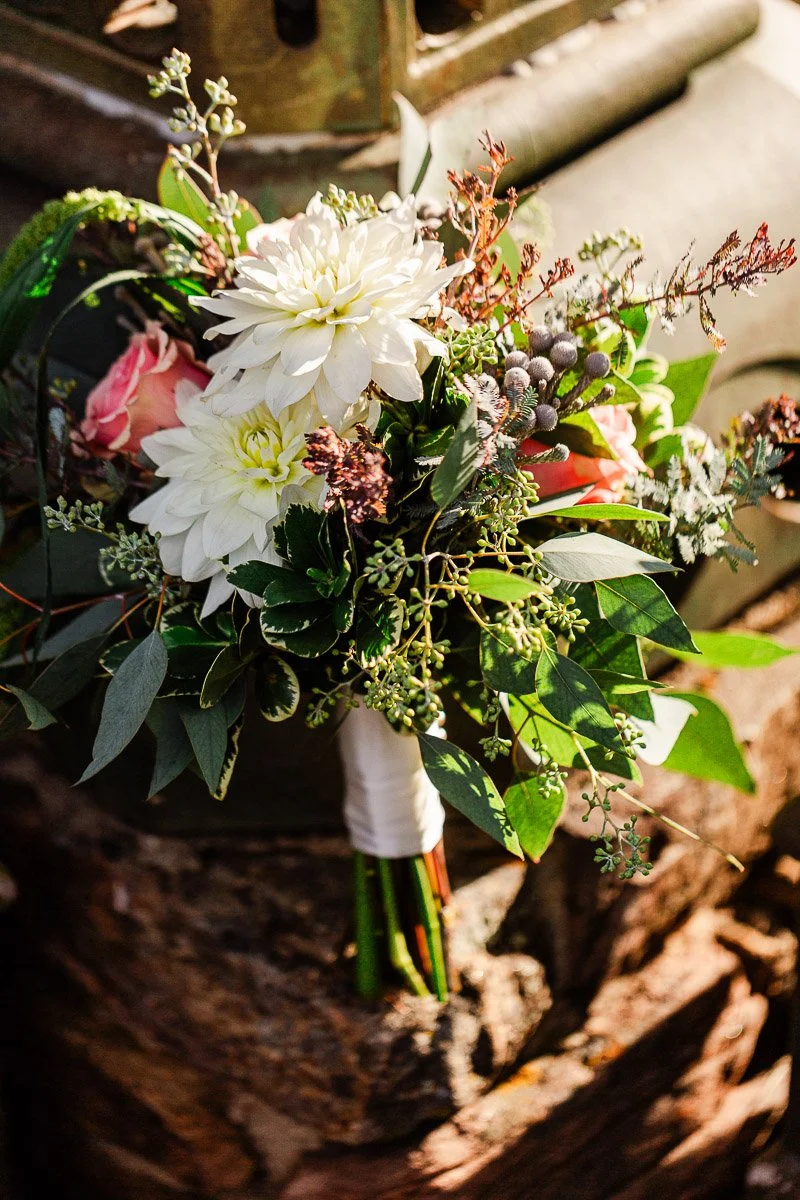 A vibrant bouquet features white dahlias, pink roses, and eucalyptus leaves. Sunlight gently highlights the rich textures and colors, creating a warm, elegant tone.