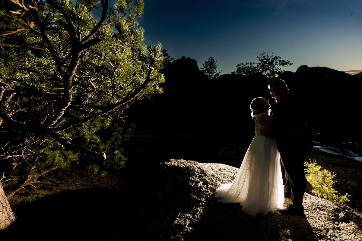Bride and groom stand on a sunlit rock at sunset, surrounded by trees. The scene is shadowed, highlighting a romantic, intimate moment.