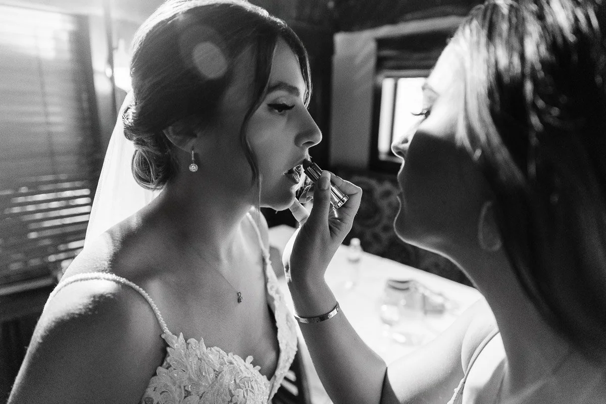 A woman in a wedding dress receives lipstick application from another woman. The black-and-white image conveys a calm, intimate, bridal preparation moment.