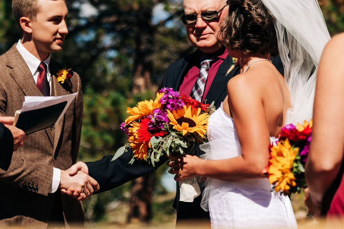 A groom in a brown suit shakes hands with an older man. The bride, in a white dress and veil, holds colorful sunflowers and blooms. Outdoor setting.