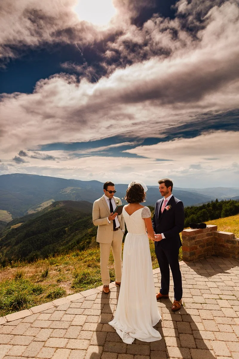 A couple stands facing each other holding hands, with a person officiating, set against a stunning mountain backdrop under a sky filled with dramatic clouds.
