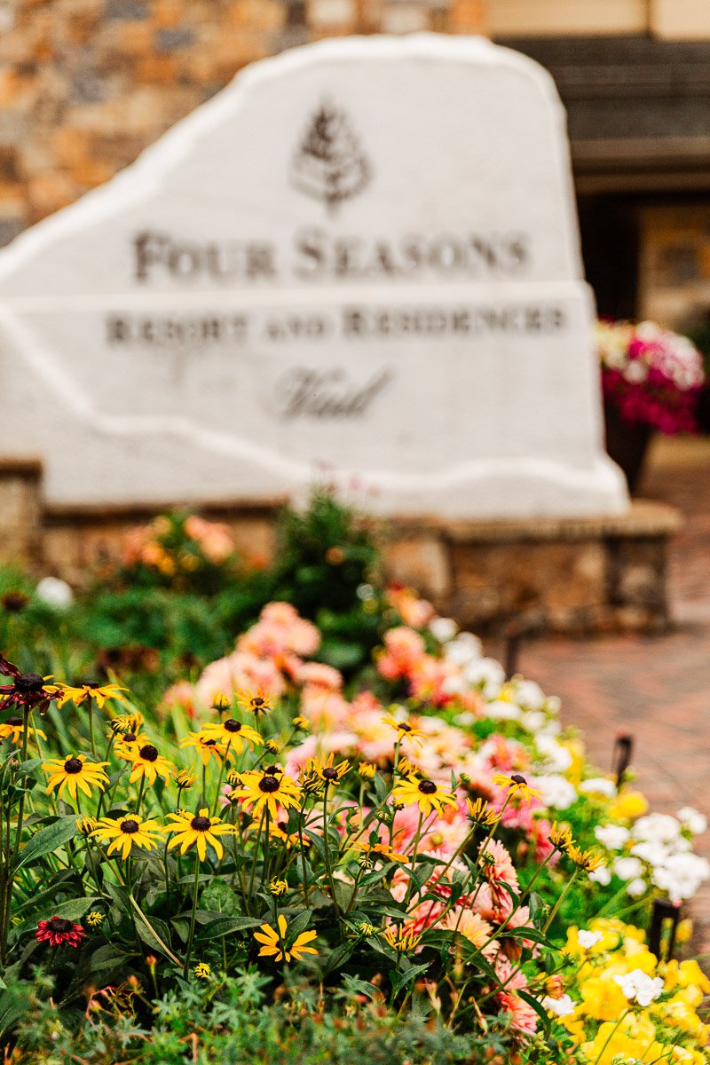 Colorful flowers, including yellow and pink blooms, in focus with a blurred stone sign in the background reading "Four Seasons Resort and Residences Vail." The scene is vibrant and welcoming.