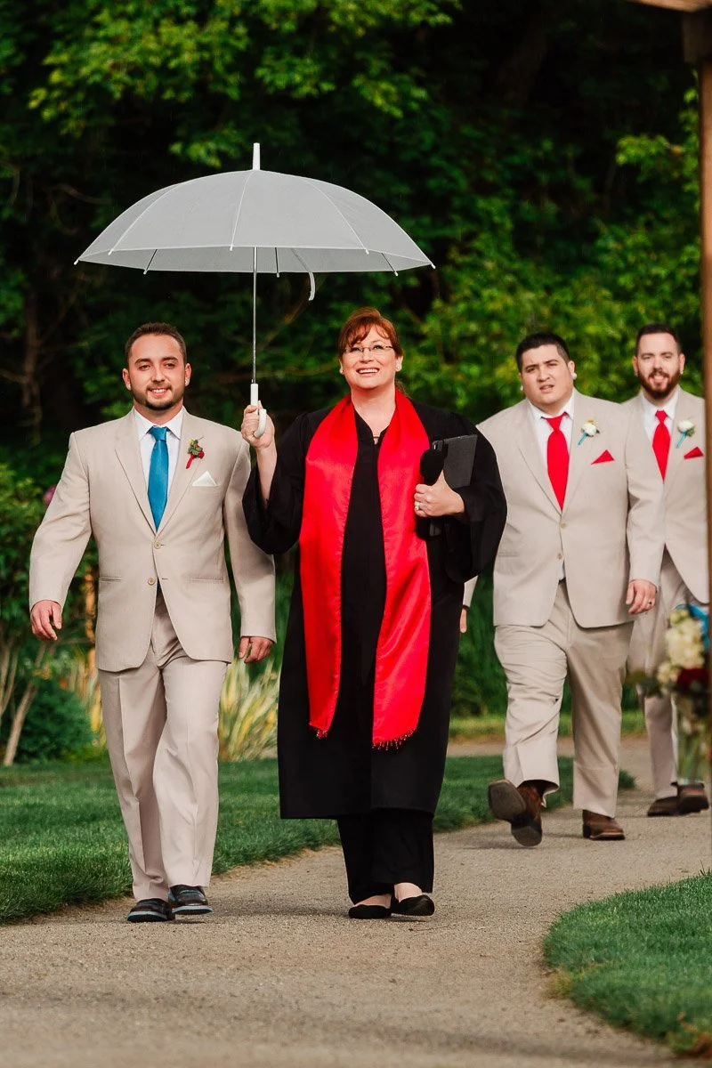 A person holding a gray umbrella walks with a smile alongside three individuals in light suits and red ties on a garden path, conveying a joyful, formal event.