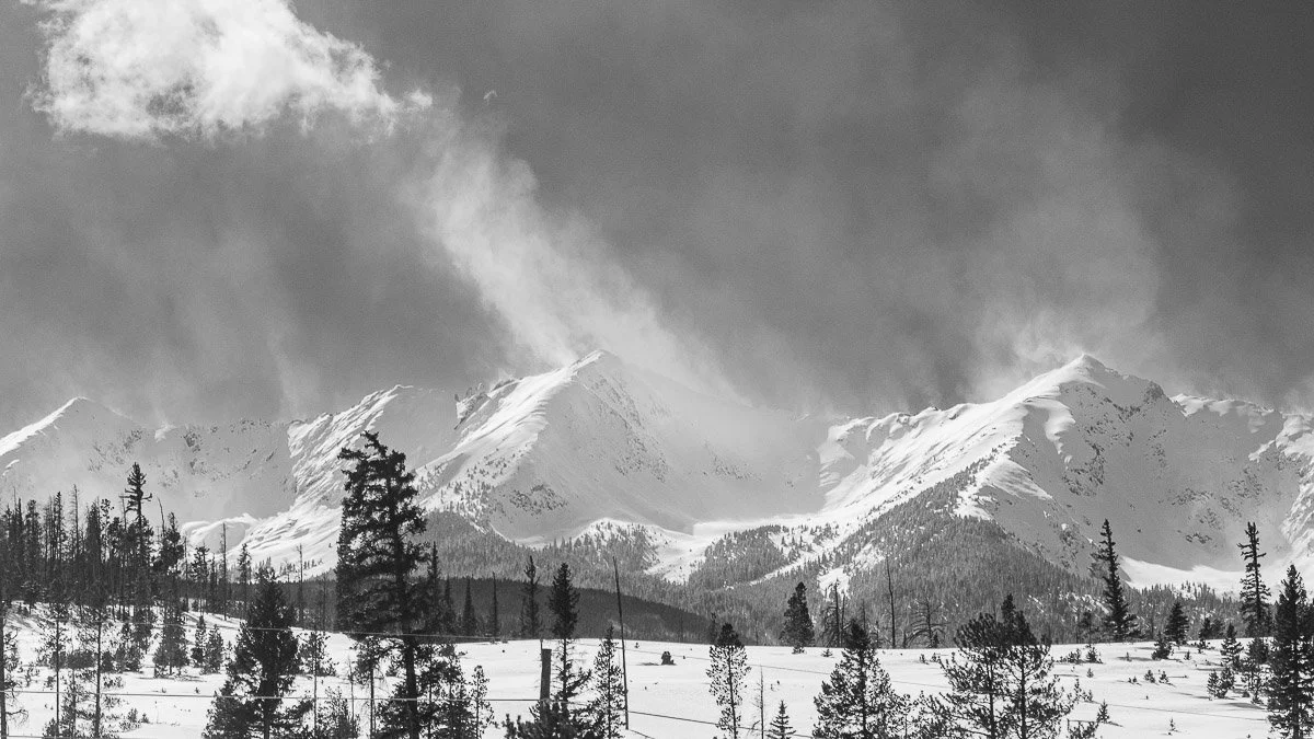 Black and white image of a snowy mountain range with dramatic clouds overhead. Tall trees in the foreground emphasize the vastness and serenity of the scene.