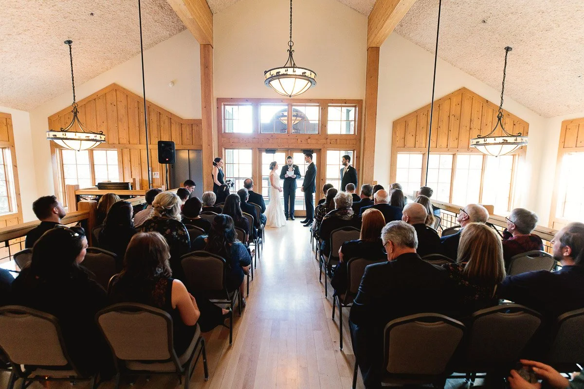 A Silverthorne Pavilion wedding ceremony in a rustic, wooden chapel. The bride and groom stand at the altar with the officiant. Guests in formal attire watch quietly.