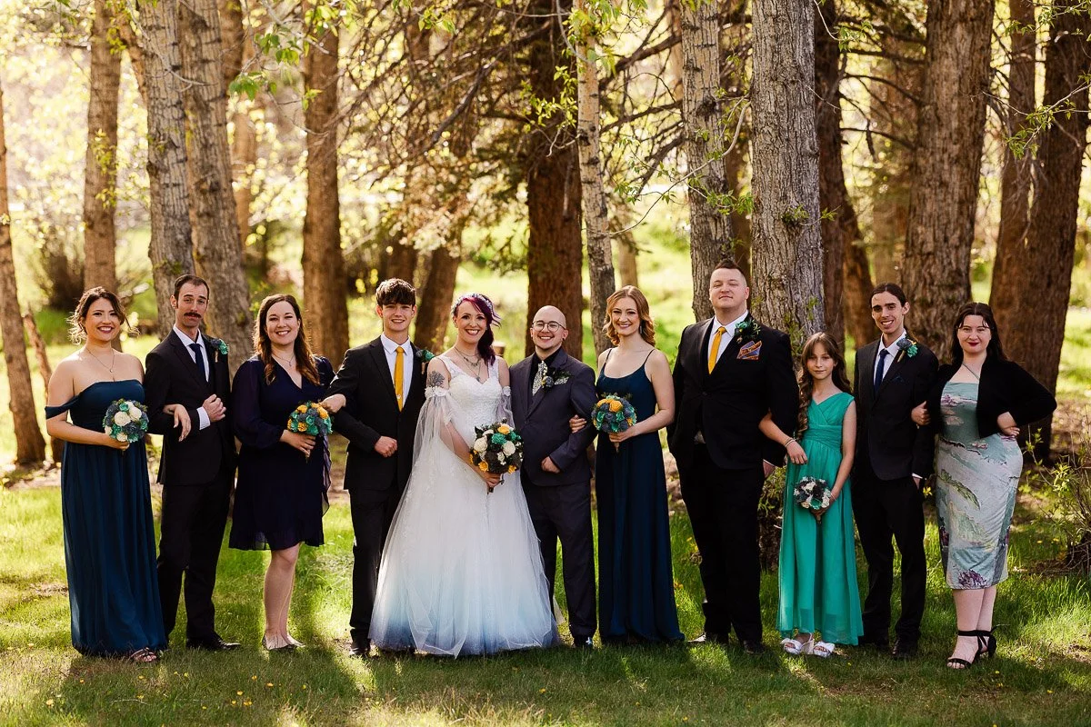 A wedding party stands in a sunlit forest. The bride in a white gown and the groom, both smiling, are surrounded by bridesmaids and groomsmen in formal attire, holding bouquets. The scene conveys joy and celebration.