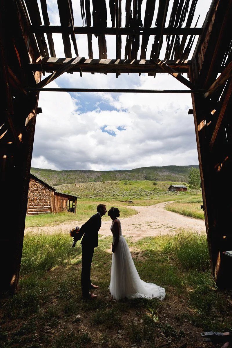 Silhouetted couple kissing beneath rustic barn entrance, bride in gown, groom with bouquet. Rolling hills and cloudy sky in background convey romance captured by Keystone wedding photographer tomKphoto