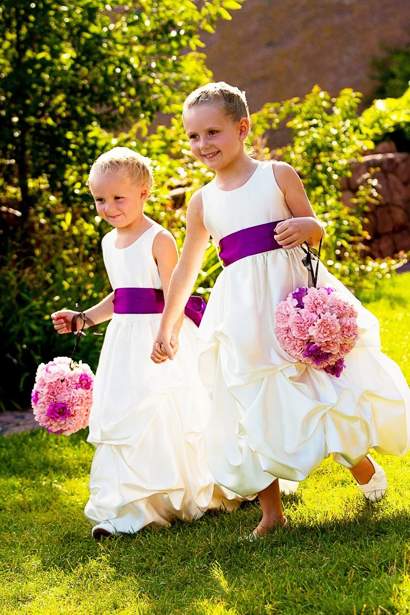 Two young girls in white dresses with purple sashes walk happily on grass, holding pink flower bouquets. The background is bright and lush with greenery.