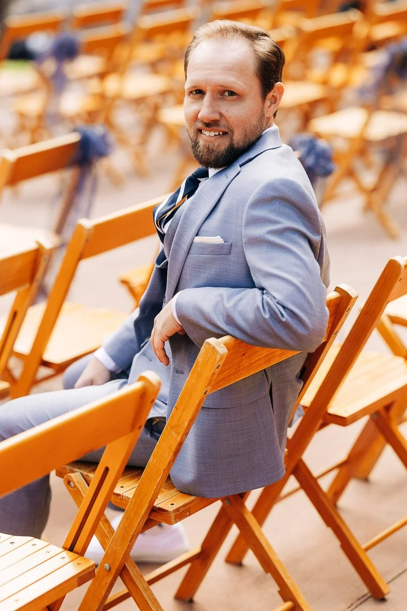 A groom in a light blue suit sits smiling on a wooden chair among rows of similar chairs, suggesting a cheerful atmosphere at an outdoor event.