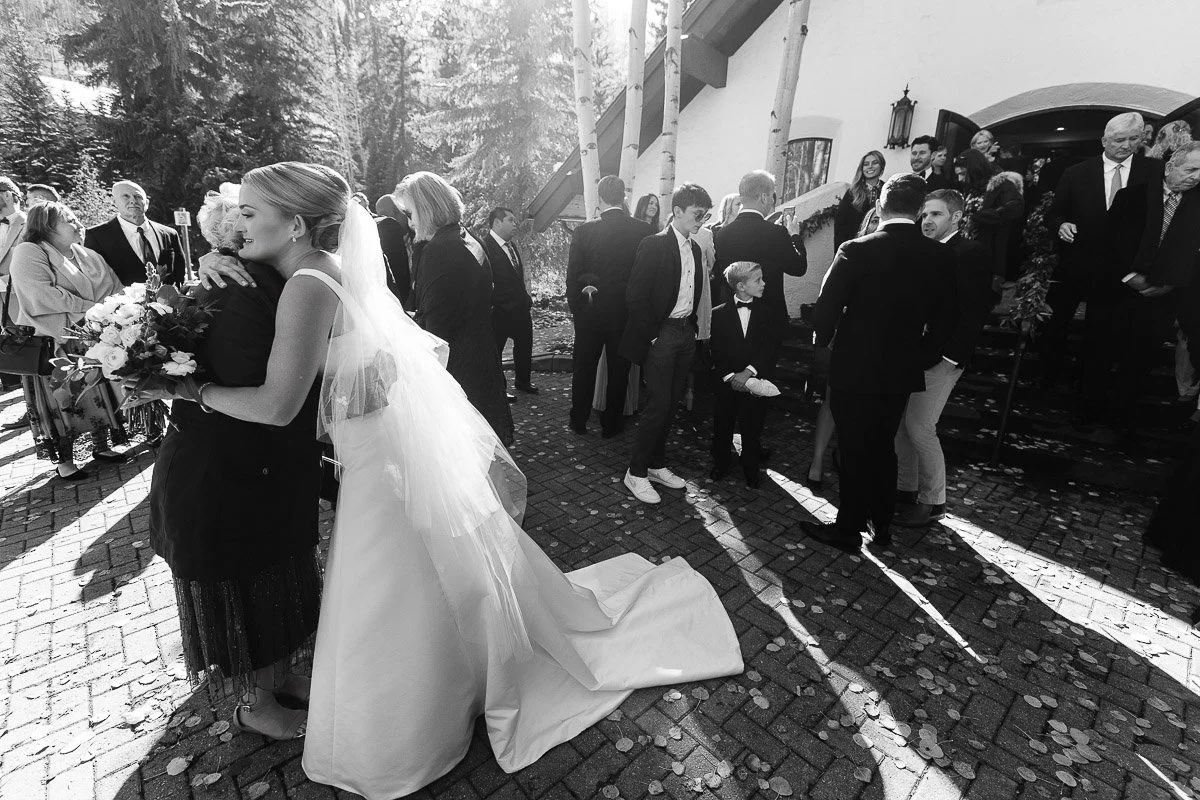 A bride in a flowing dress and veil embraces a guest outdoors, surrounded by people in formal attire. The scene is joyful and lively with sunlight filtering through trees.