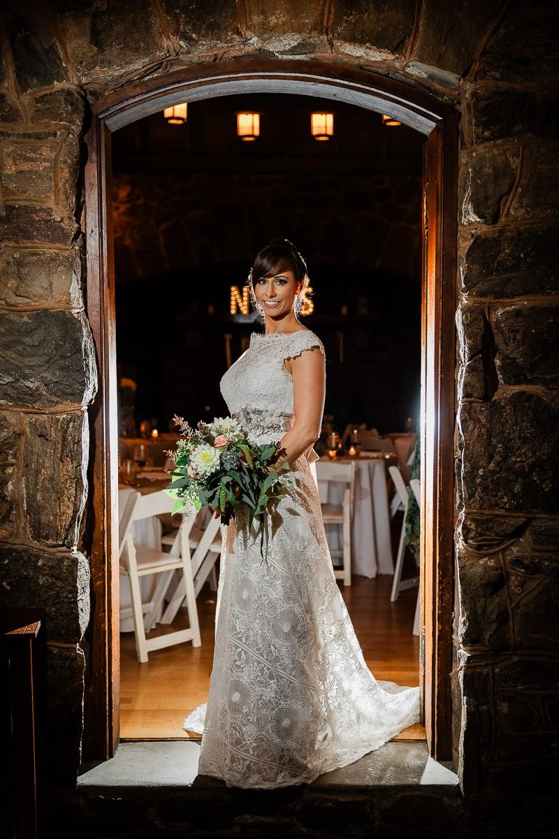 A bride in a lace gown and veil stands in a stone archway, holding a bouquet of colorful flowers. Warm light creates an elegant, joyful ambiance.